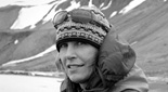 Photo of Elizabeth Bradfield in a winter parka and knitted cap, beside a shoreline with snowy hills in the background.